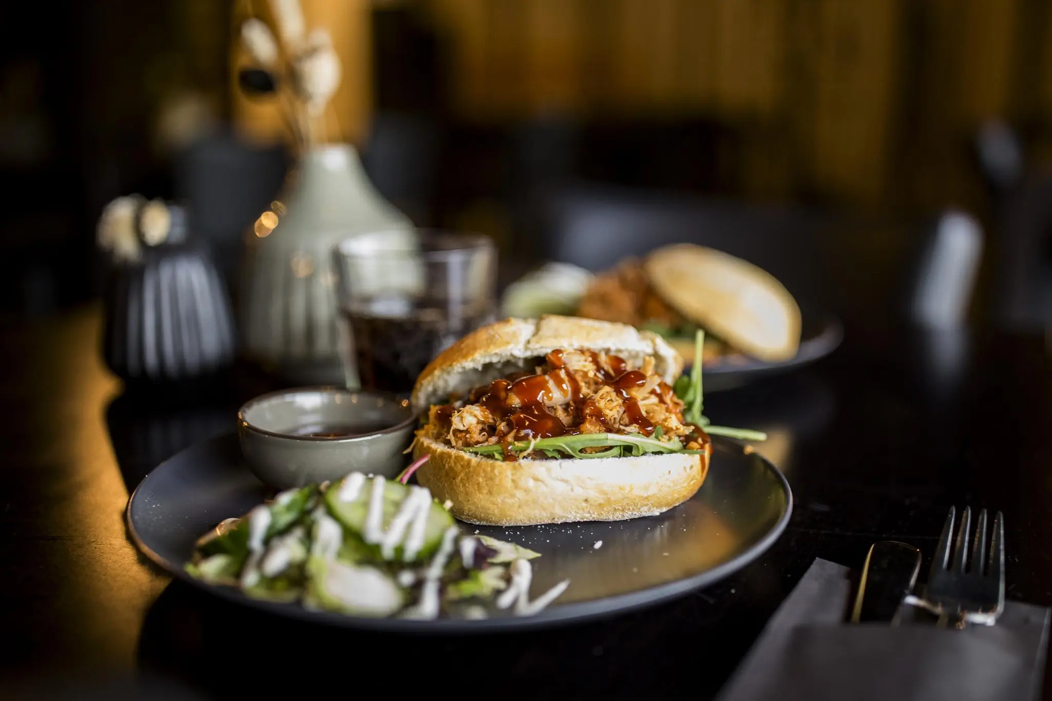 Broodje met pulled chicken, barbecuesaus en sla geserveerd met een frisse salade en dipsaus op een donker bord in restaurant Markerkant 13.